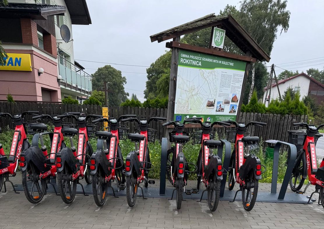 9 rowerów zaparkowanych na parkingu rowerowym. Rowery są w kolorze czarno-czerwonym. W tle widać tablicę z napisem Rokitnica i blok mieszkalny w kolorze biało-różowym.